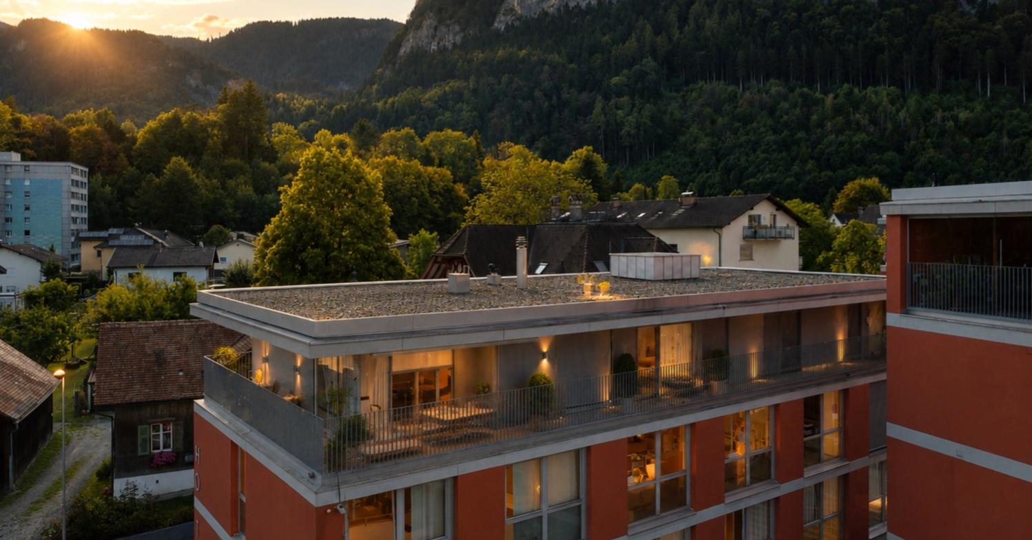Penthouse mit beleuchteter Dachterrasse in der Abenddämmerung vor bewaldeten Berghängen